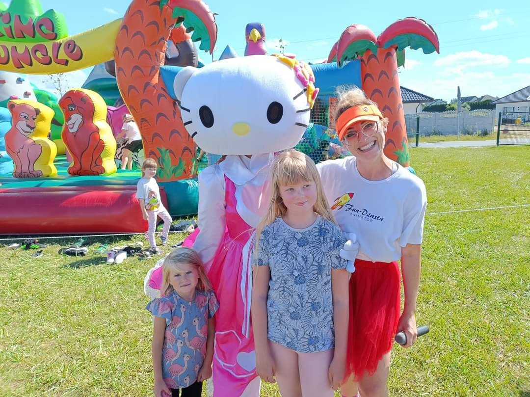 Uśmiechnięta kobieta w pomarańczowym daszku i kostium Hello Kitty pozują z dwiema dziewczynkami na tle dmuchanego placu zabaw z motywem dżungli.