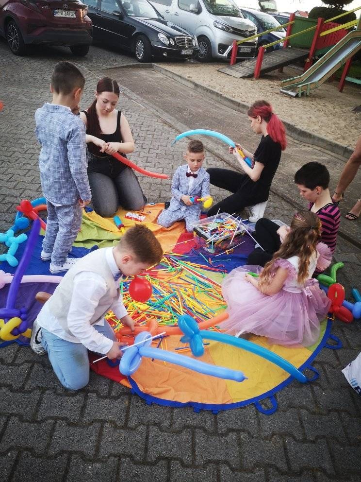 Grupa dzieci i dwie animatorki siedzą na kolorowej macie na zewnątrz, tworząc figury z balonów. Dzieci ubrane w eleganckie stroje, a animatorki w czarnych ubraniach. W tle widoczne samochody i plac...