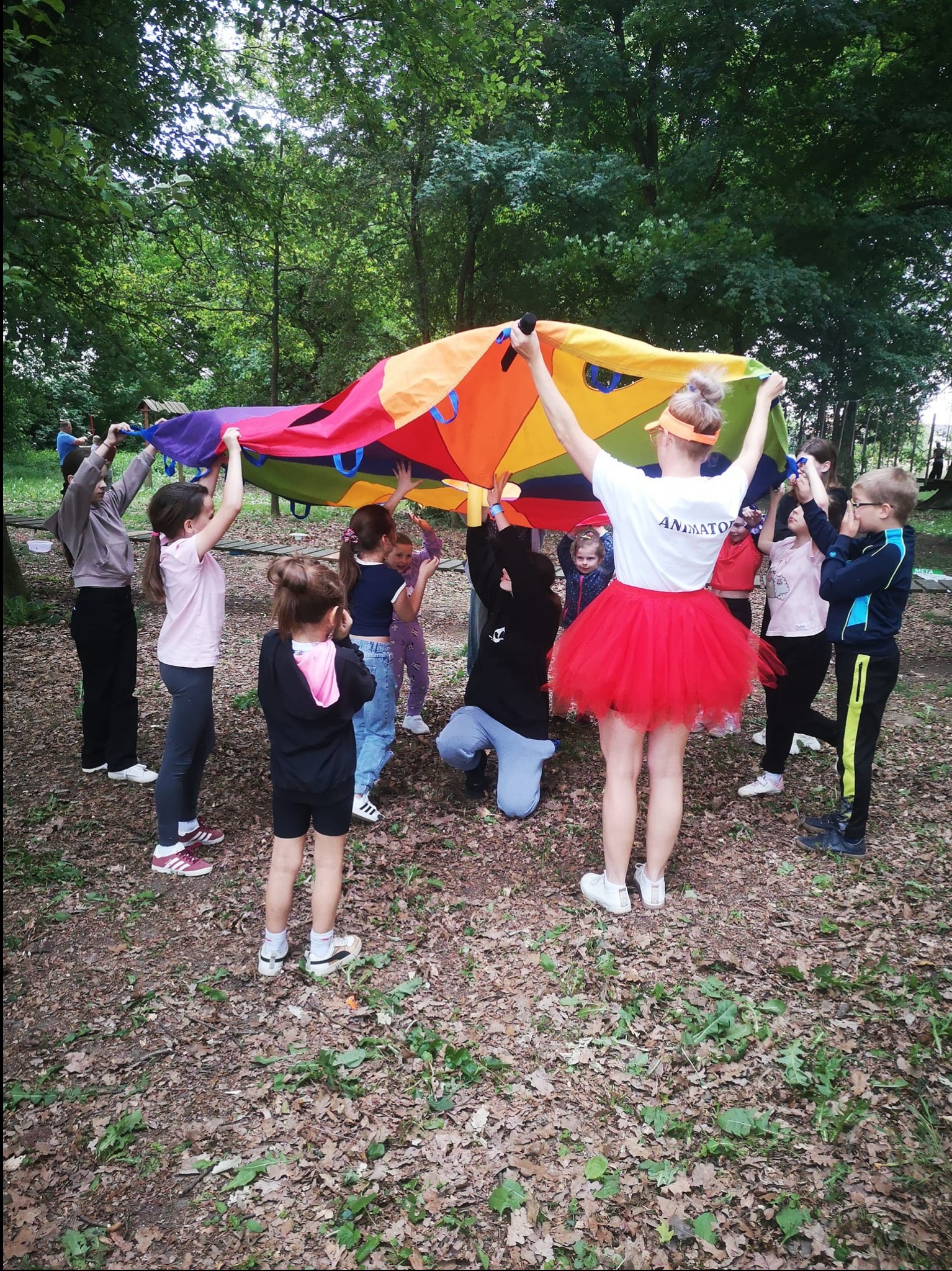 Grupa dzieci trzymająca kolorową chustę animacyjną pod nadzorem kobiety w czerwonej tiulowej spódnicy i koszulce z napisem, zabawa na świeżym powietrzu w otoczeniu drzew.