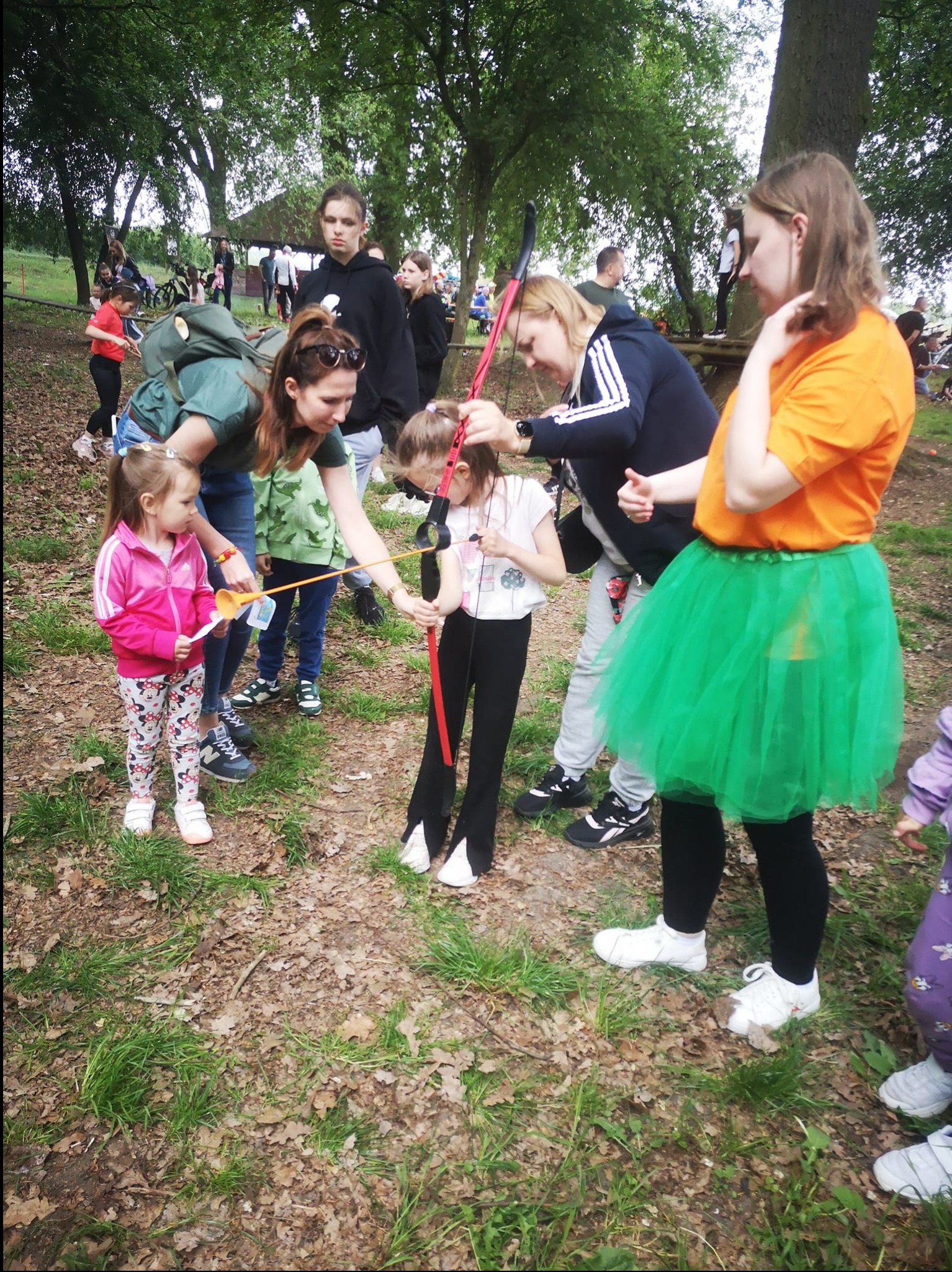 Dziewczynka uczy się strzelać z łuku pod okiem instruktorów w plenerze, w tle widoczni inni uczestnicy zabawy.