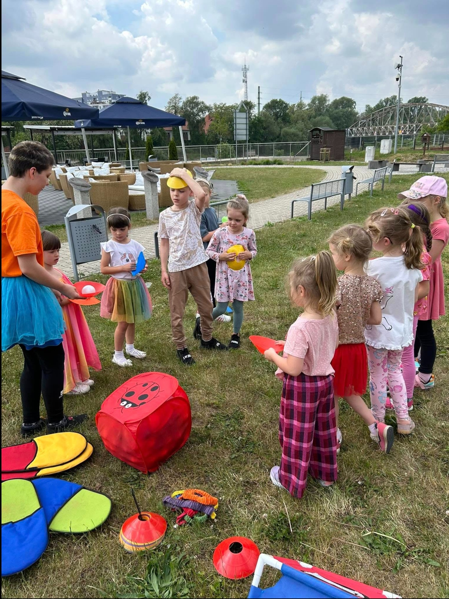 Animator w pomarańczowej koszulce prowadzi zabawy z grupą dzieci na trawie, używając rekwizytów takich jak talerzyki, piłki i duża kostka z biedronką, w tle widoczny teren z parasolami i budynkami.