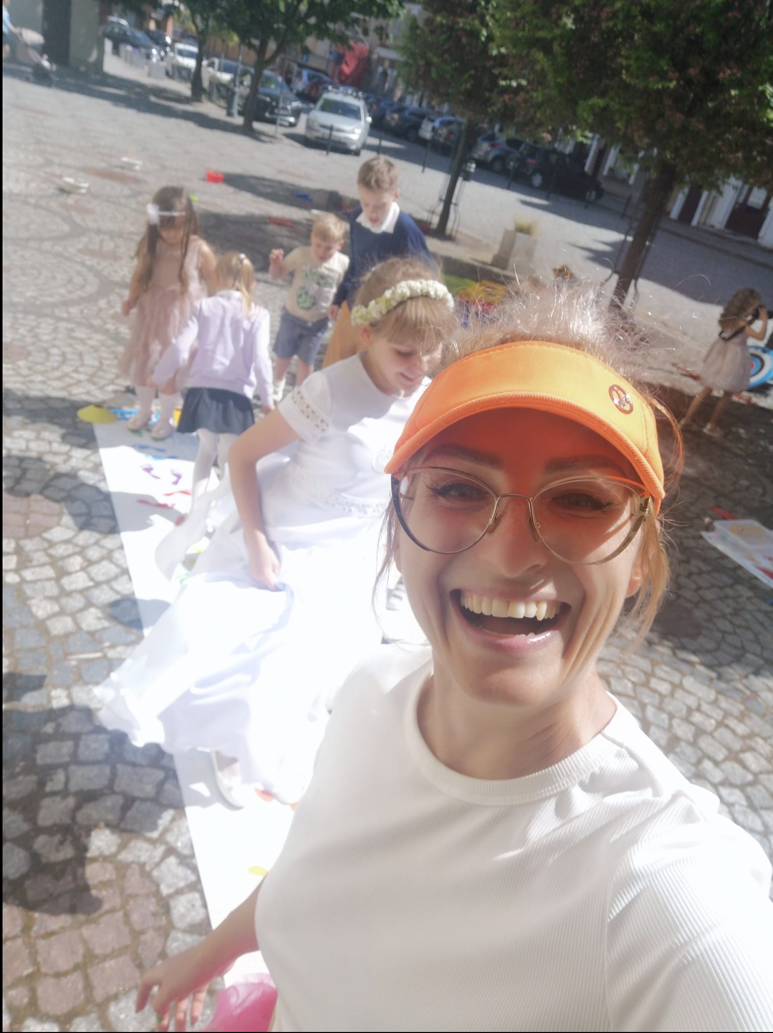 Uśmiechnięta kobieta w pomarańczowym daszku robi selfie na tle dzieci grających w grę na chodniku podczas imprezy plenerowej.