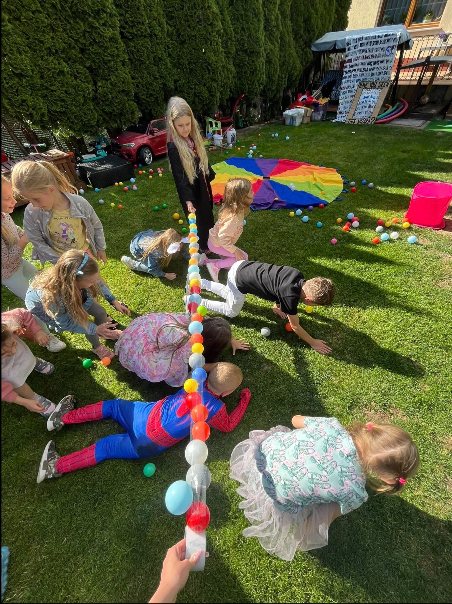 Grupa dzieci na trawie bierze udział w zabawie z kolorowymi piłeczkami w tubie, jedno dziecko przebrane za Spidermana leży na ziemi, w tle widoczny ogród z dmuchanym zamkiem i czerwonym samochodem.
