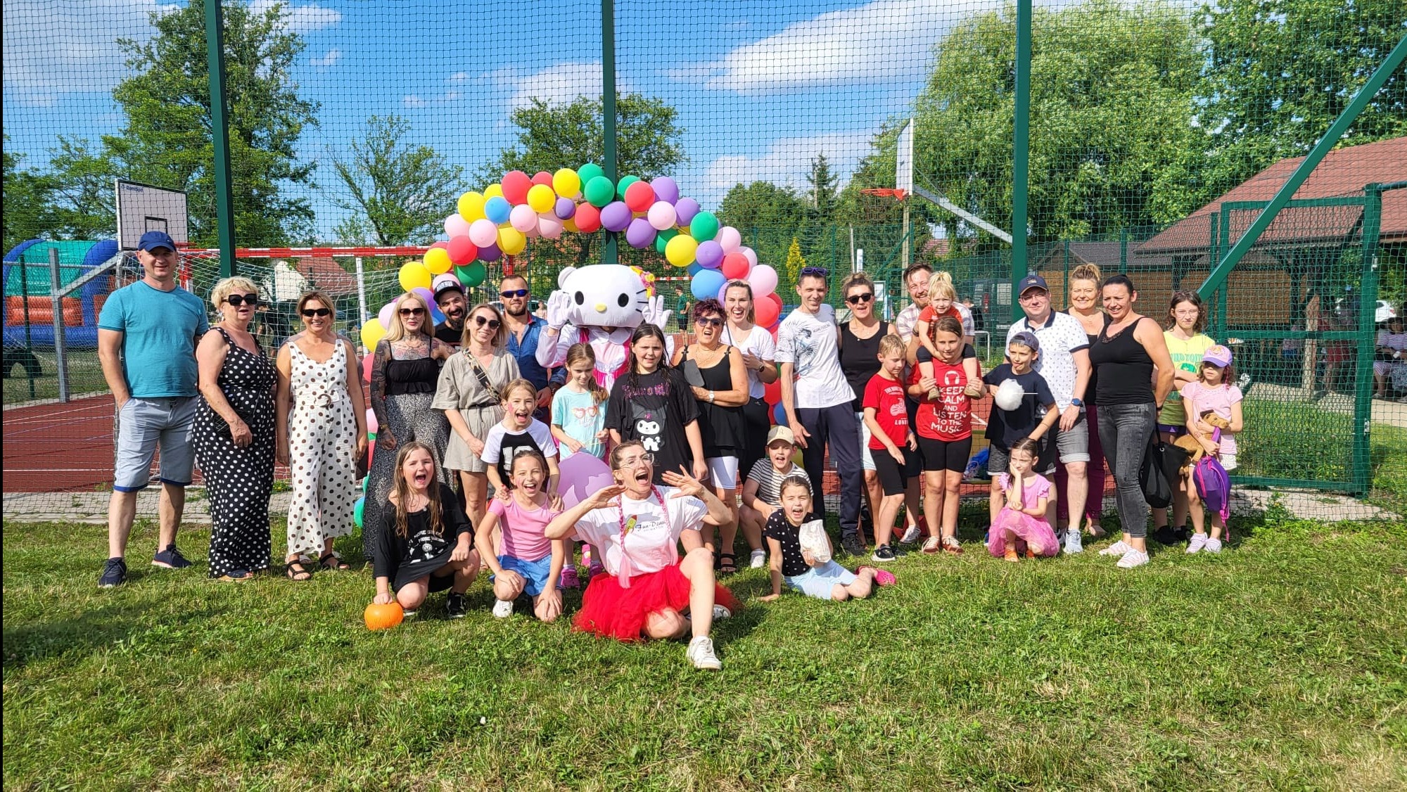 Duża grupa ludzi, w tym dzieci, pozuje do zdjęcia na trawie przed dmuchanym łukiem z balonów i postacią z bajki w kształcie kota. W tle widoczna siatka ogrodzeniowa i elementy boiska.