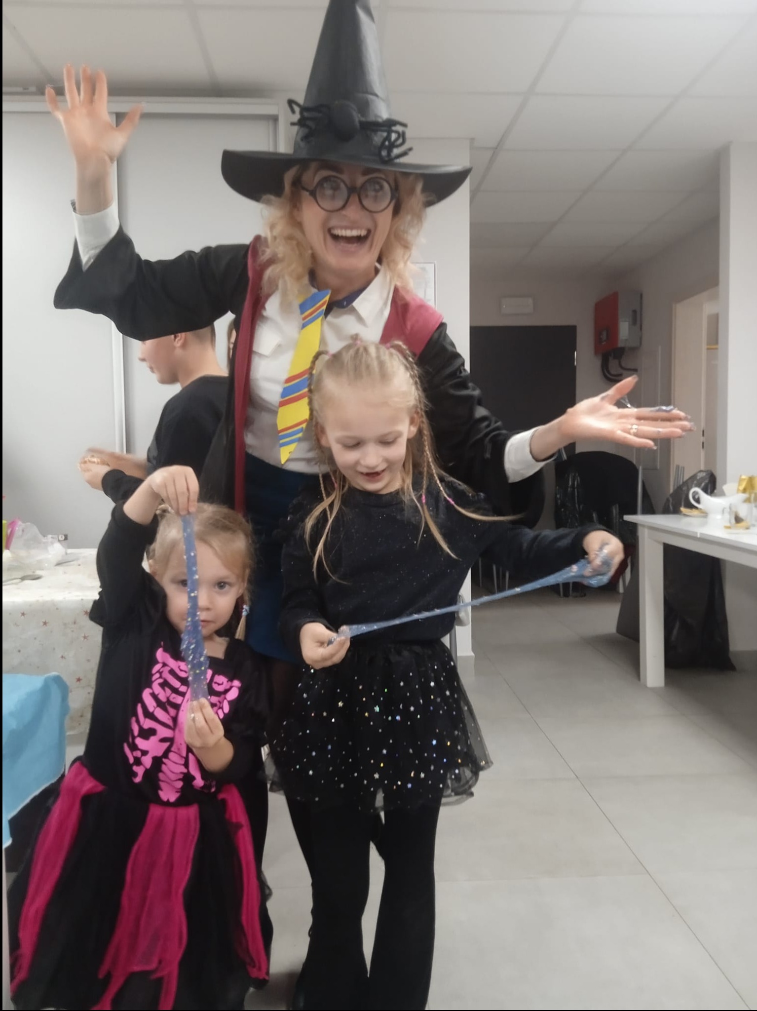 Uśmiechnięta kobieta w czarodziejskim kapeluszu i stroju, przypominającym postać z Harry'ego Pottera, bawi się z dwiema dziewczynkami, które trzymają w rękach błyszczący slime. Jedna z dziewczynek...