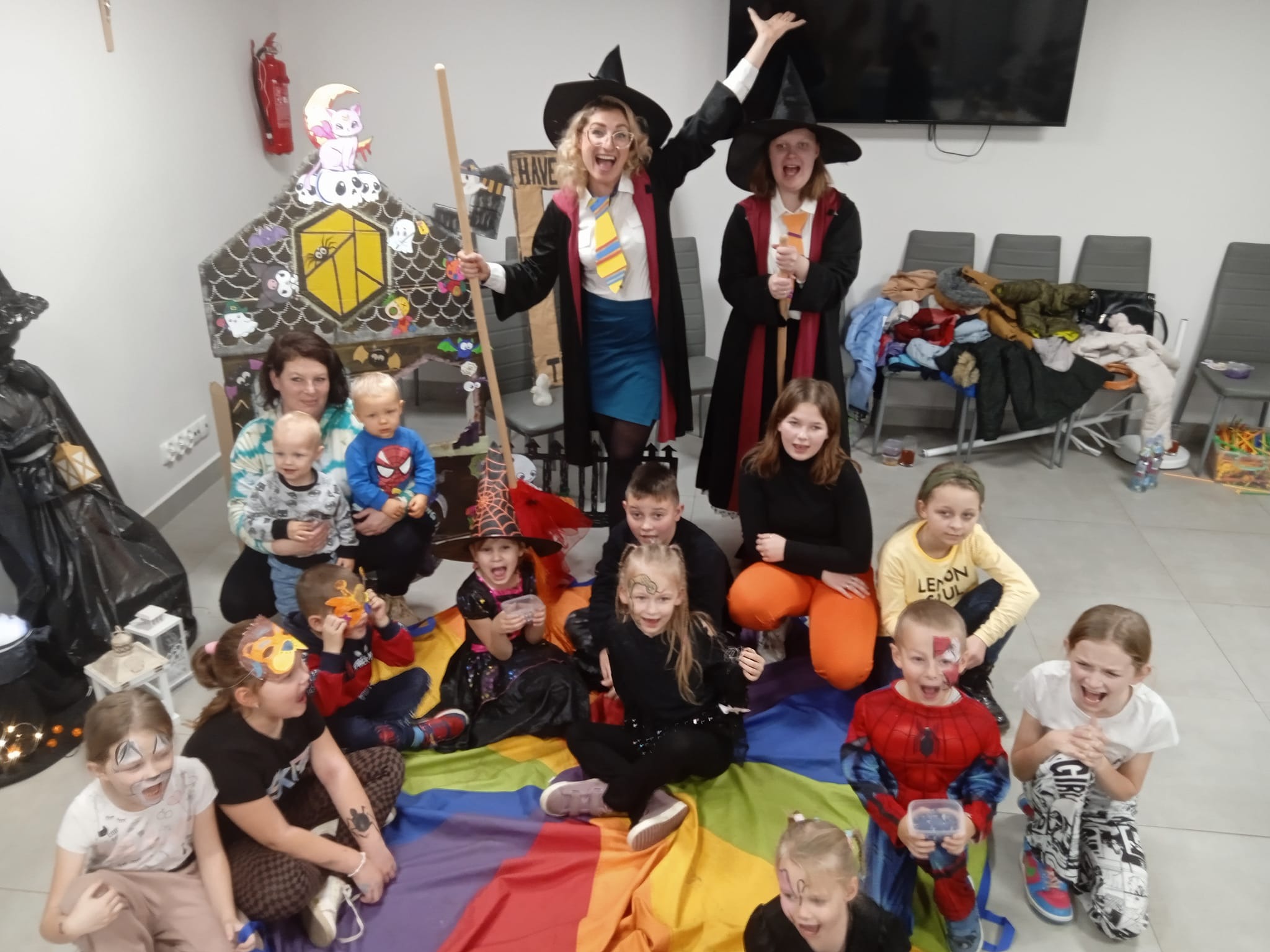 Grupa dzieci w kostiumach, w tym dwie osoby przebrane za postacie z Harry'ego Pottera, pozuje do zdjęcia na tęczowej płachcie podczas imprezy tematycznej. W tle dekoracje i dorośli.