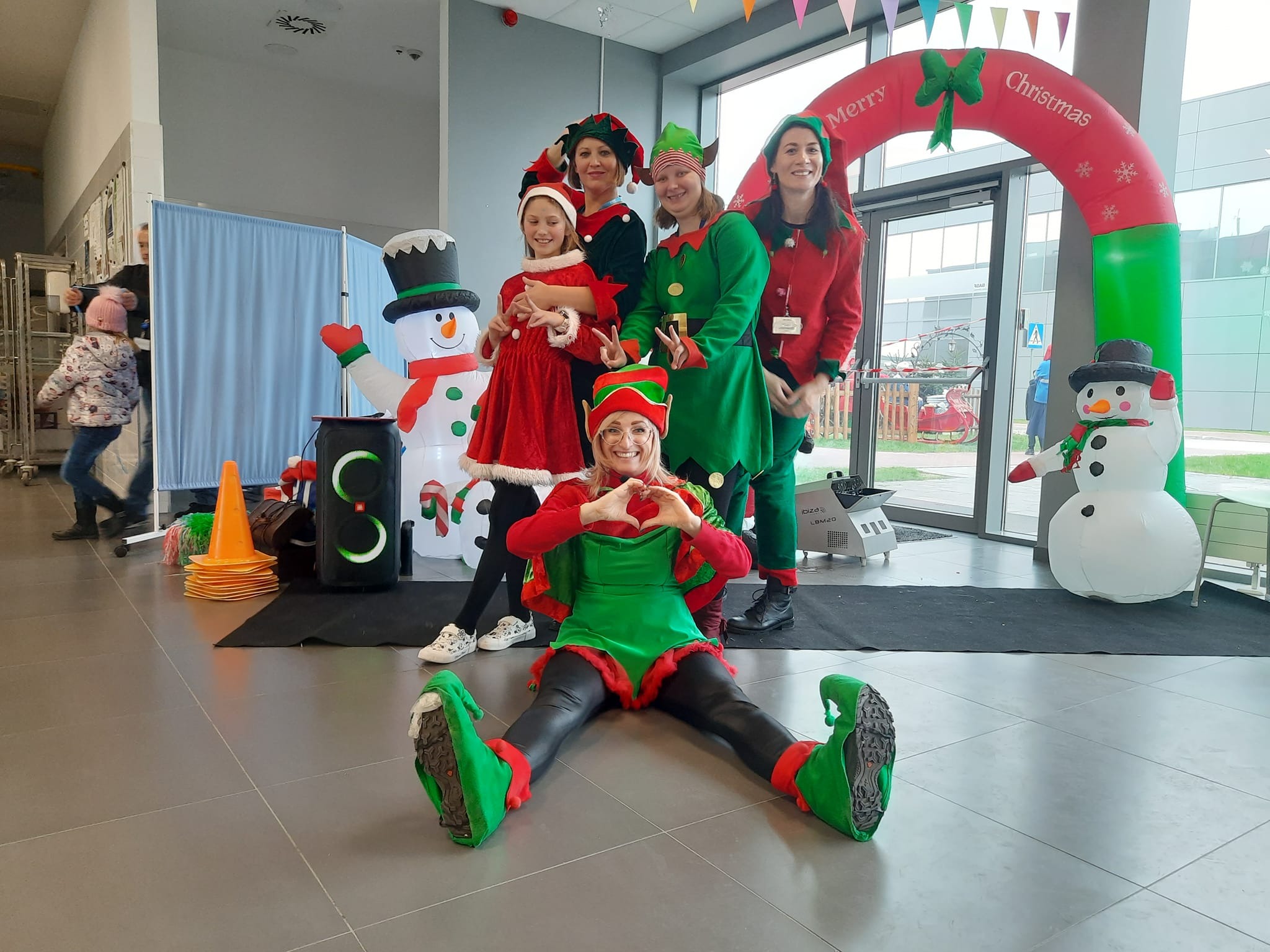 Grupa animatorów przebranych za elfy w świątecznych strojach pozuje do zdjęcia w pomieszczeniu z dmuchanym łukiem 'Merry Christmas' i dmuchanymi bałwanami, jedna z elfek siedzi na podłodze, tworząc...