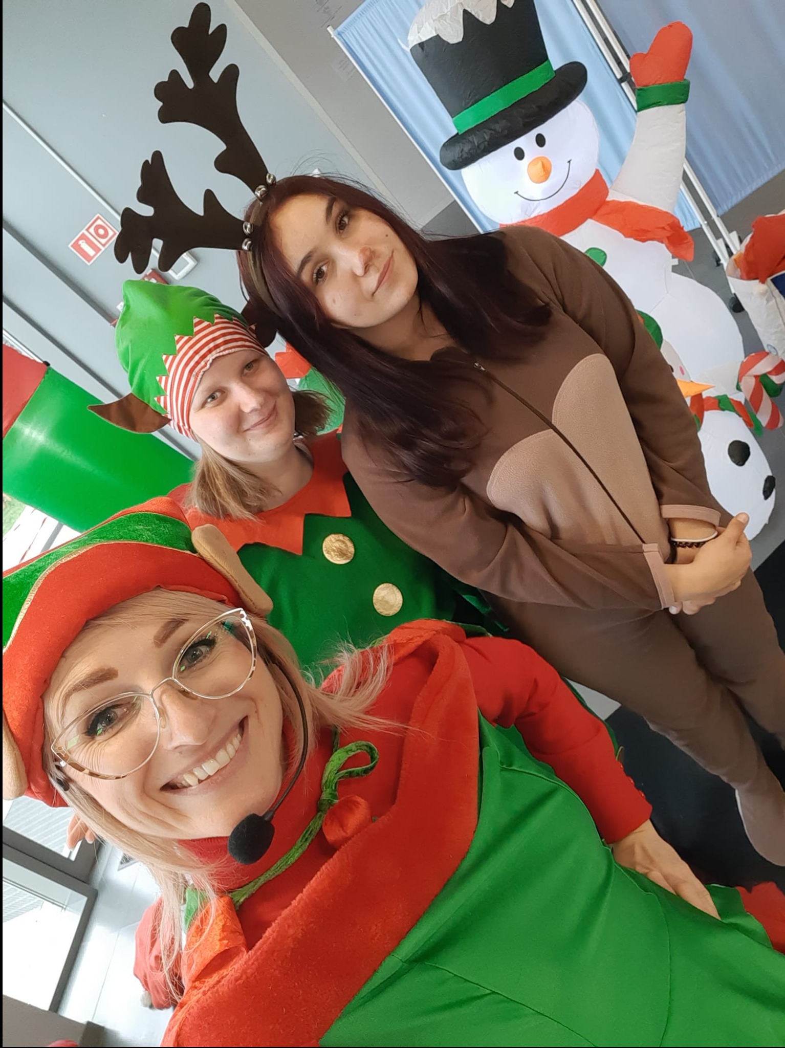 Trzy osoby w kostiumach: dwie przebrane za elfy w czerwono-zielonych strojach i czapkach, jedna w brązowym kombinezonie renifera z rogami, pozują do selfie na tle dmuchanego bałwana.