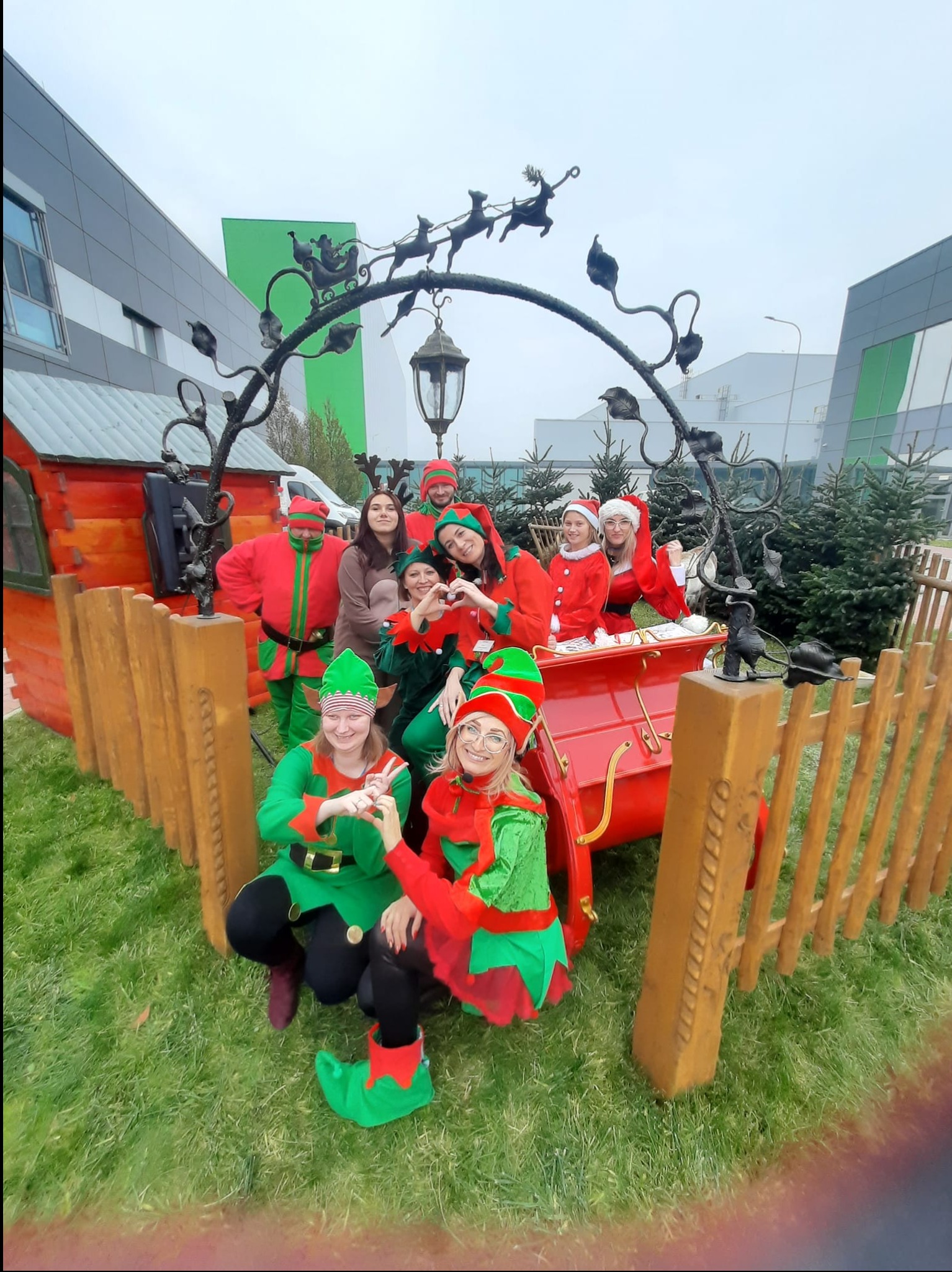 Grupa osób przebranych za elfy i Mikołaja pozuje do zdjęcia obok czerwonych sań z dekoracją reniferów i domku z drewna na tle nowoczesnego budynku.
