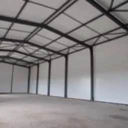 Wnętrze nowo wybudowanej hali z widoczną stalową konstrukcją nośną i białymi ścianami z płyt. Widoczne łączenia płyt na ścianach. Hala jest pusta, przygotowana do dalszego użytkowania.