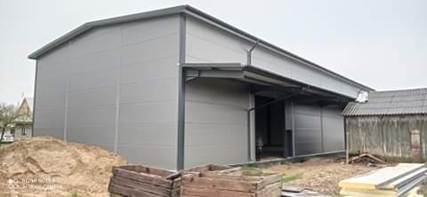 Szara hala magazynowa z otwartymi przesuwnymi drzwiami, widoczny stos piasku i drewniane skrzynie na pierwszym planie, zarośnięty budynek gospodarczy w tle.