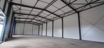 Wnętrze nowo wybudowanej hali z widoczną stalową konstrukcją nośną i białymi ścianami z płyt. Widoczne łączenia płyt na ścianach. Hala jest pusta, przygotowana do dalszego użytkowania.