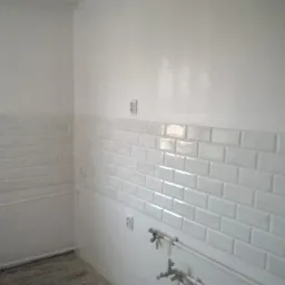 Świeżo położone, białe, prostokątne płytki ceramiczne na ścianie w pomieszczeniu, widoczne gniazdka elektryczne i rury instalacji wodnej, ściana nad płytkami pomalowana na biało.