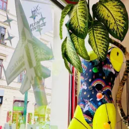 Wystawa sklepowa z manekinem w kostiumie z motywem jednorożców, rośliną doniczkową i naklejką samolotu na szybie z napisem 'Flyin' Vintage', widok na ulicę.