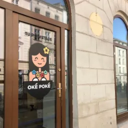 Okleina na drzwiach lokalu 'OKÉ POKÉ' z grafiką dziewczyny z kwiatami i napisem 'TU POWSTAJE' w odbiciu szyby