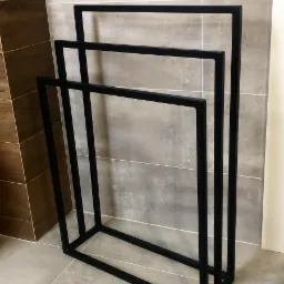 Czarny, metalowy stojak na ręczniki łazienkowe z trzema poziomami, wykonany ze spawanych profili o kwadratowym przekroju, na tle szarej i brązowej glazury.