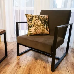 Nowoczesny fotel z ciemnoszarą tapicerką i czarną, metalową ramą, ozdobiony poduszką w tropikalne liście, stojący na drewnianej podłodze w jasnym wnętrzu z zasłonami.