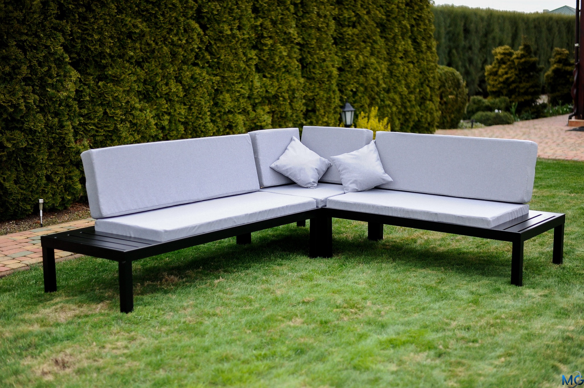 Kątowy zestaw mebli ogrodowych z czarną metalową ramą i szarymi poduszkami na trawie, z żywopłotem w tle.
