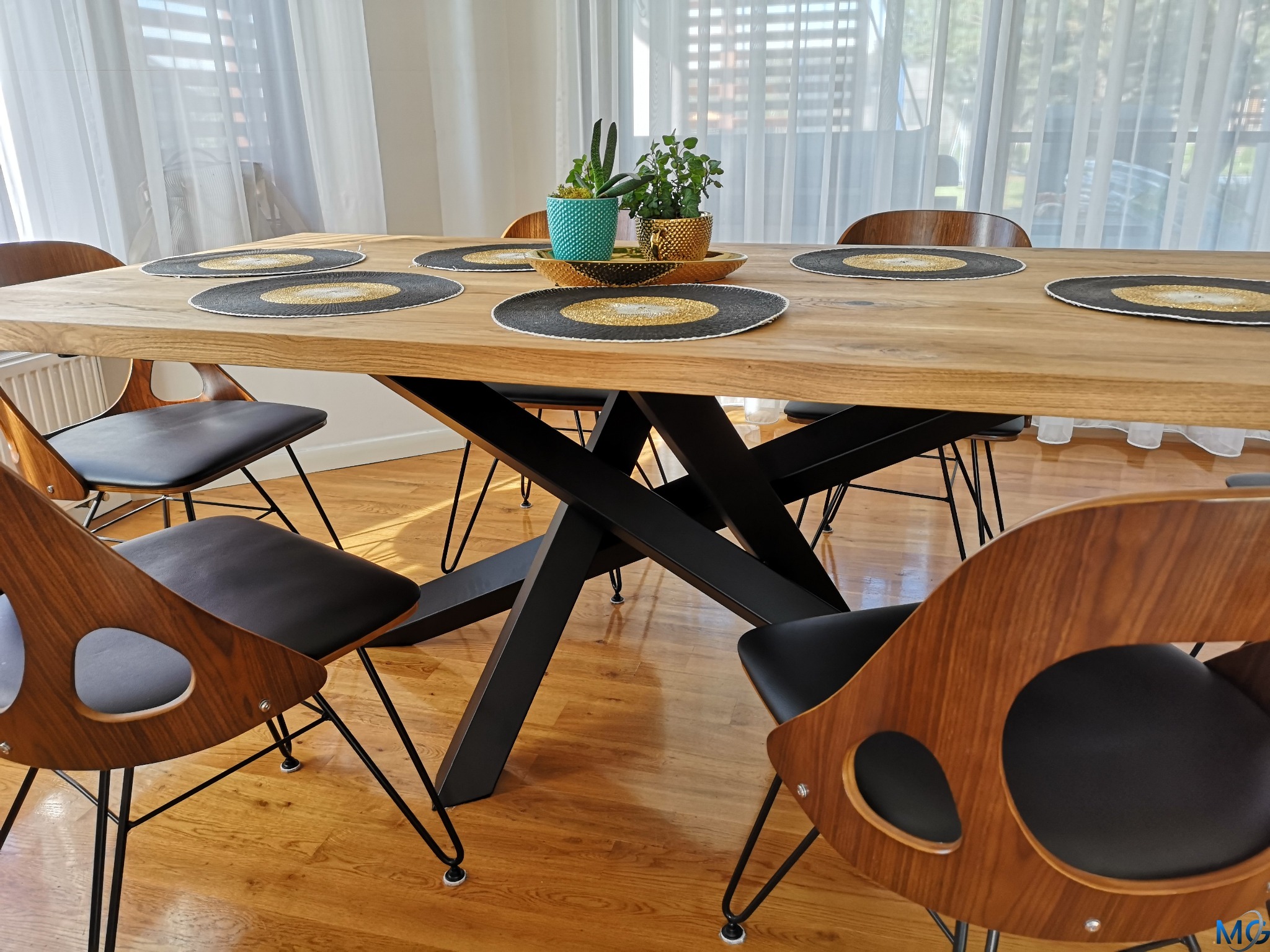Stół z blatem z jasnego drewna i czarnymi metalowymi nogami w kształcie litery X, otoczony krzesłami z drewnianymi oparciami i czarnymi siedziskami, na stole dekoracyjne podkładki i rośliny...