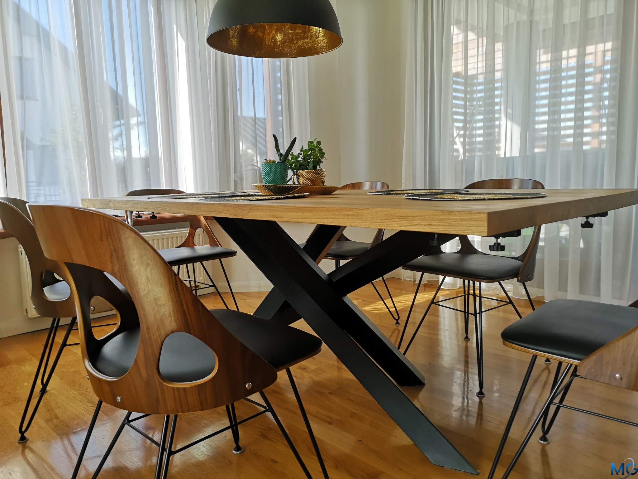 Stół z blatem z jasnego drewna i czarną, metalową podstawą w kształcie litery X, otoczony krzesłami z giętego drewna i czarnymi siedziskami, całość oświetlona złotą lampą wiszącą i naturalnym...