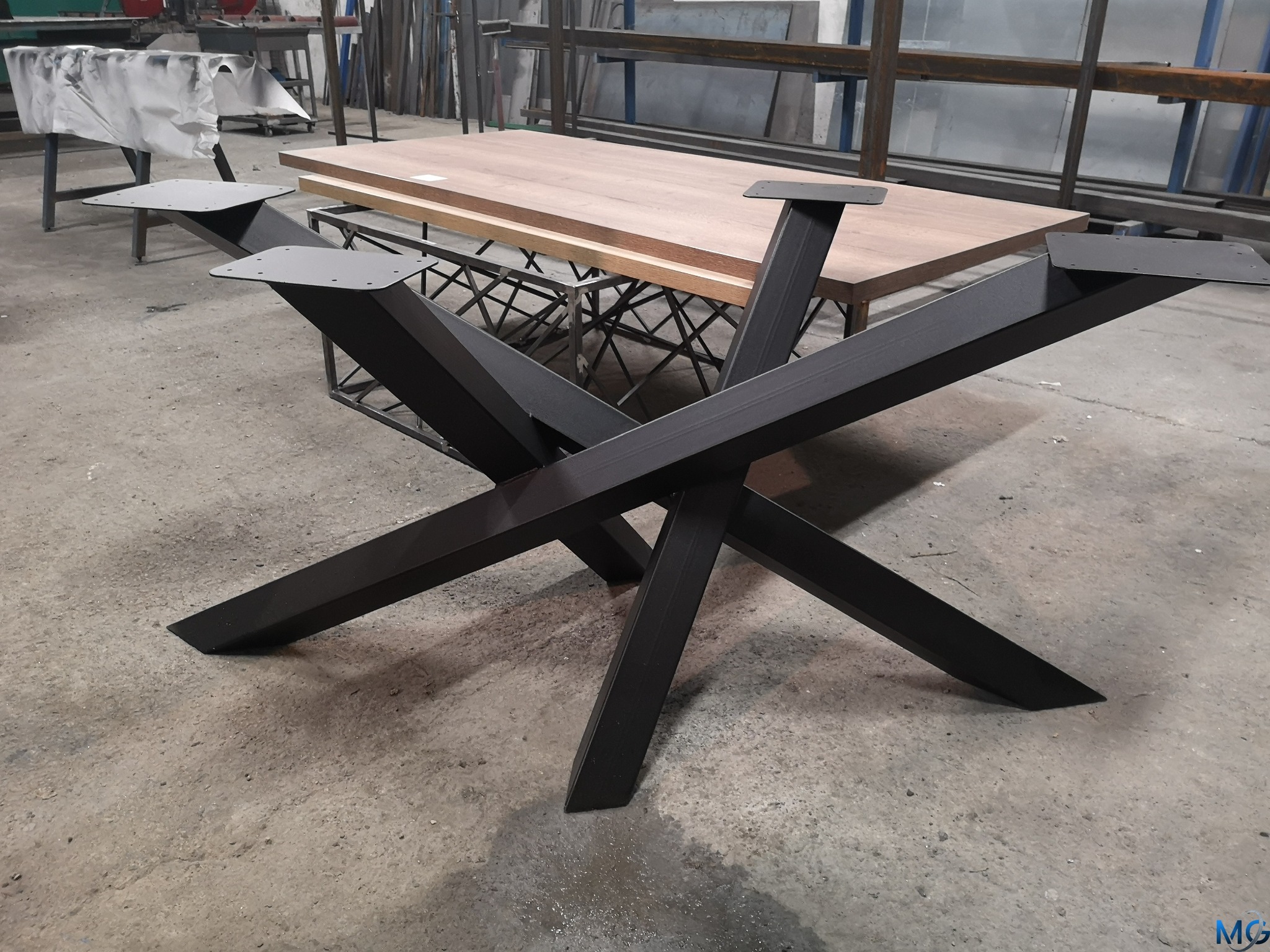 Czarny, metalowy stelaż stołu z geometrycznym układem nóg, widoczny na tle warsztatu z elementami konstrukcji stalowych i blatem stołu z drewna.