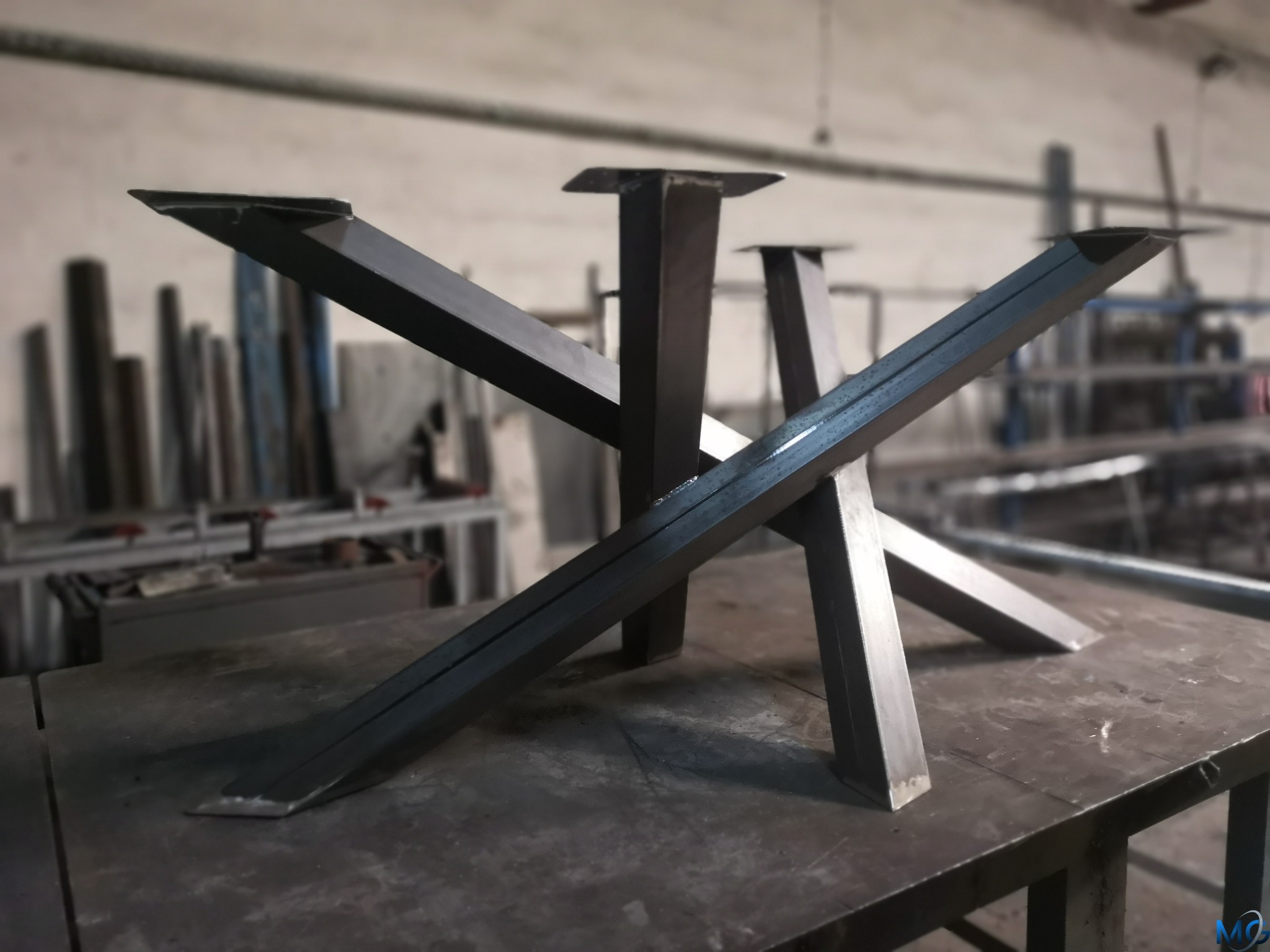 Metalowe nogi do stołu w kształcie litery X, spawane z profili stalowych, surowe wykończenie, widoczne spawy, tło warsztatowe z elementami konstrukcji stalowych.