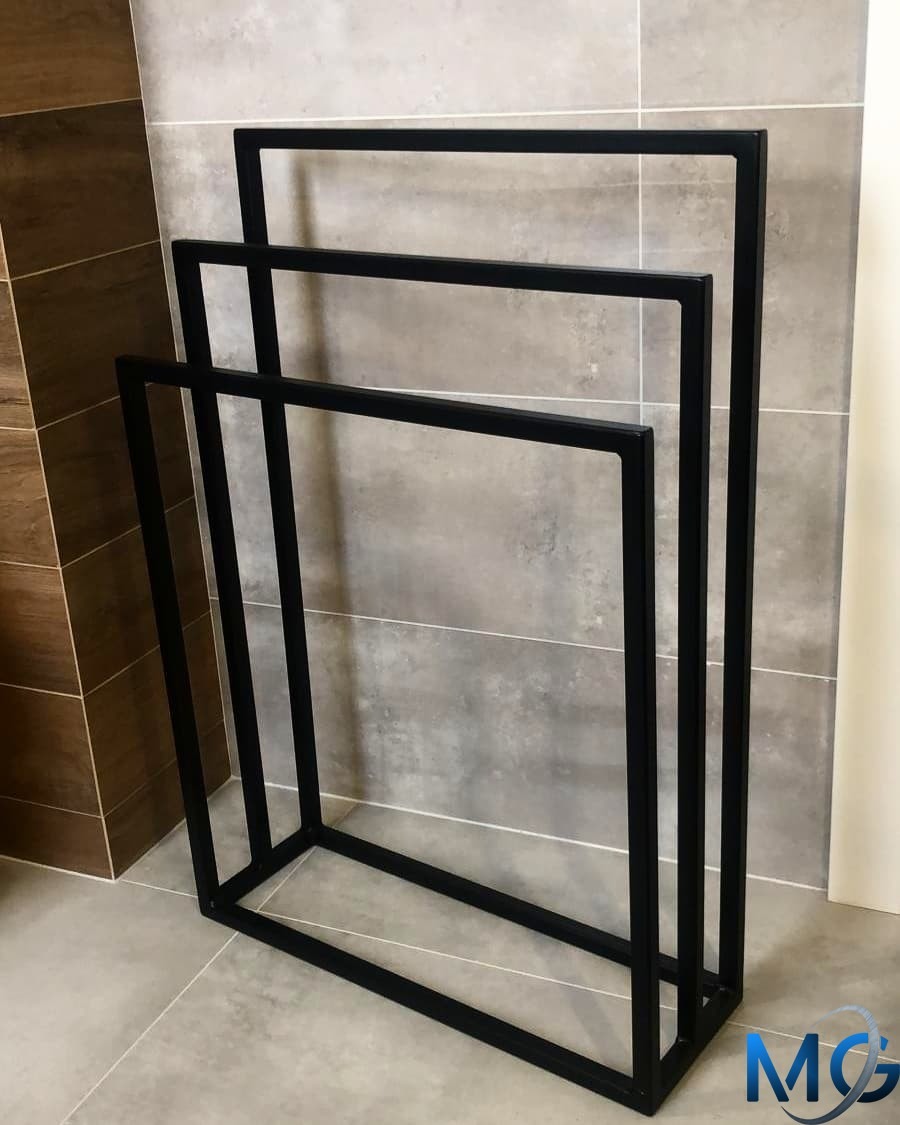 Czarny, metalowy stojak na ręczniki łazienkowe z trzema poziomami, wykonany ze spawanych profili o kwadratowym przekroju, na tle szarej i brązowej glazury.