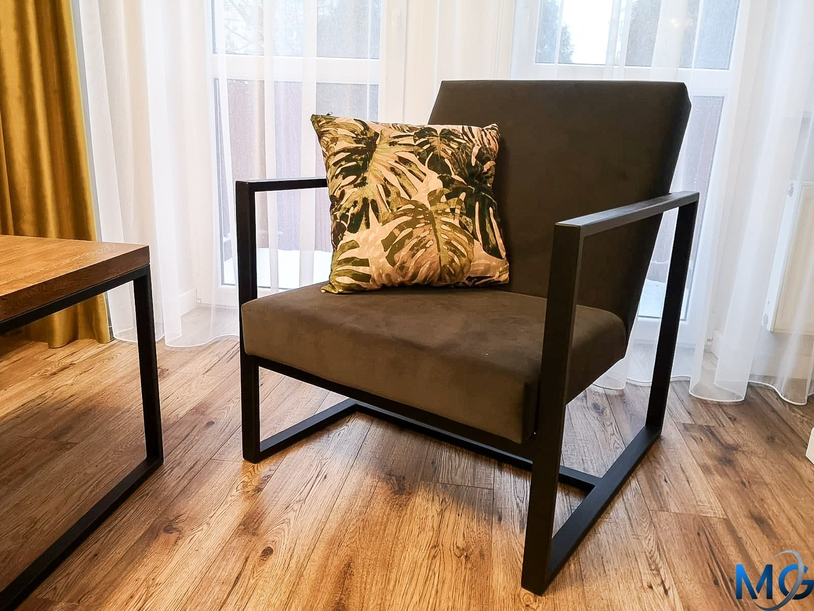Nowoczesny fotel z ciemnoszarą tapicerką i czarną, metalową ramą, ozdobiony poduszką w tropikalne liście, stojący na drewnianej podłodze w jasnym wnętrzu z zasłonami.