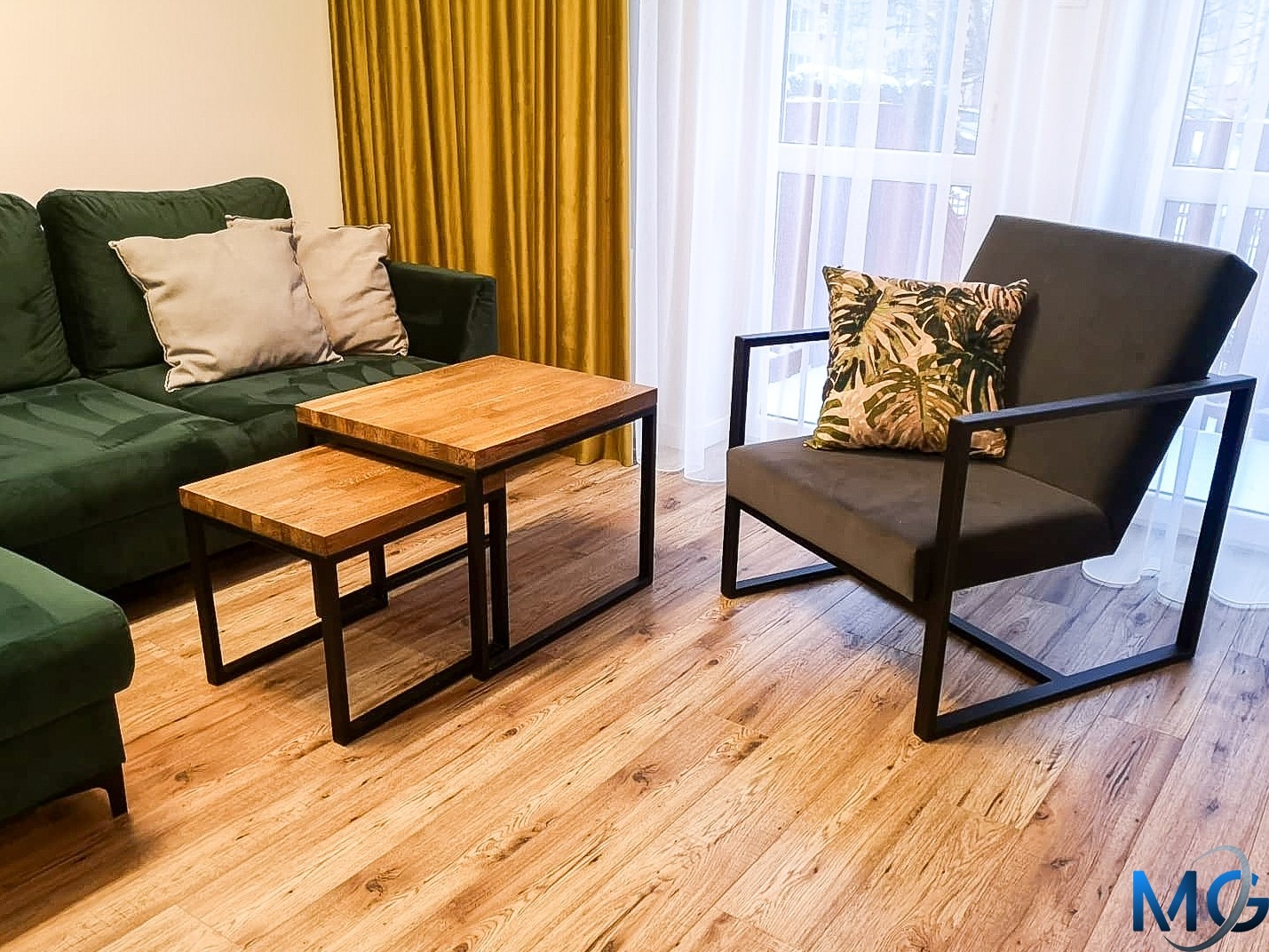 Nowoczesny salon z zieloną sofą, drewnianymi stolikami kawowymi z czarnymi metalowymi nogami oraz szarym fotelem z poduszką w liście na drewnianej podłodze.