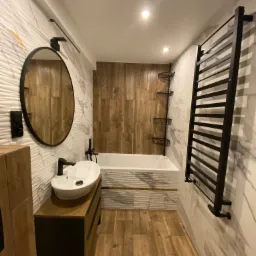 Nowoczesna łazienka po remoncie: drewnopodobne płytki, owalne lustro, biała umywalka nablatowa i czarny grzejnik drabinkowy. Wykończenie wnętrz w ciepłych barwach.