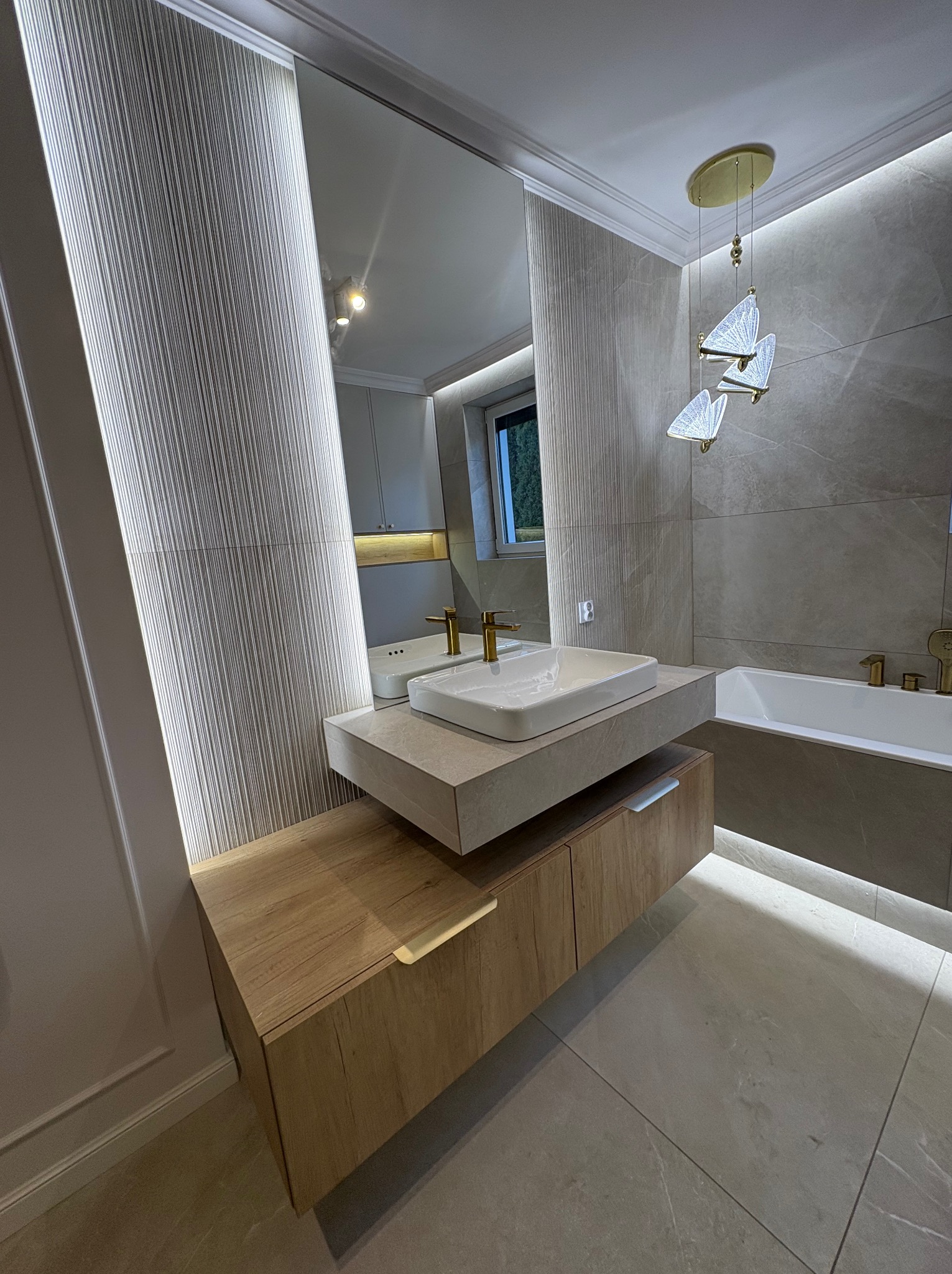 Nowoczesna łazienka z podświetlanym lustrem i umywalką na szafce z drewnianym frontem. Na suficie designerska lampa w kształcie motyli. Szare płytki na ścianach i podłodze.