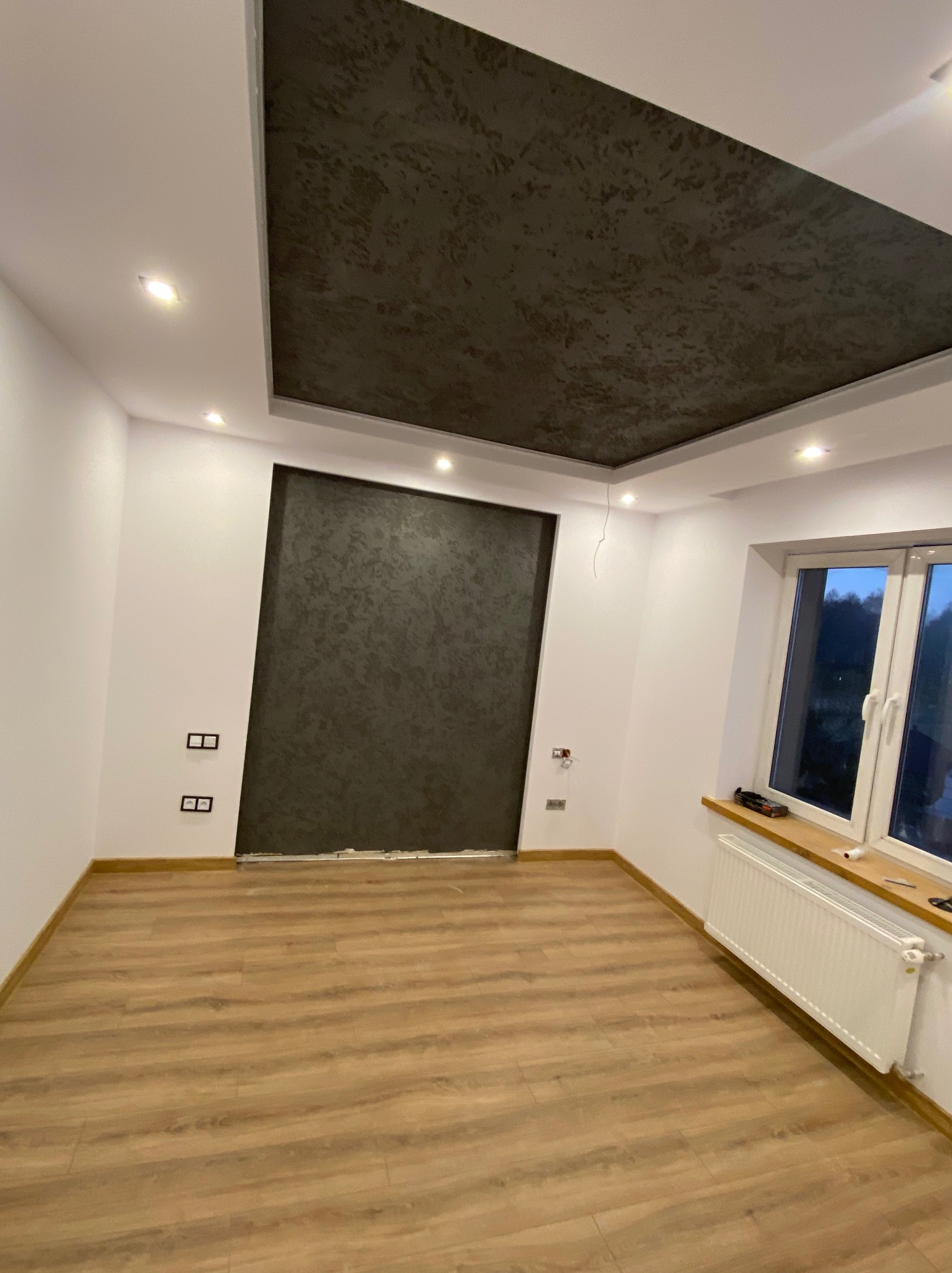 Nowoczesne wykończenie wnętrza: parkiet, białe ściany, dekoracyjny sufit podwieszany z szarą strukturą. Widoczne gniazdka, okno z grzejnikiem.