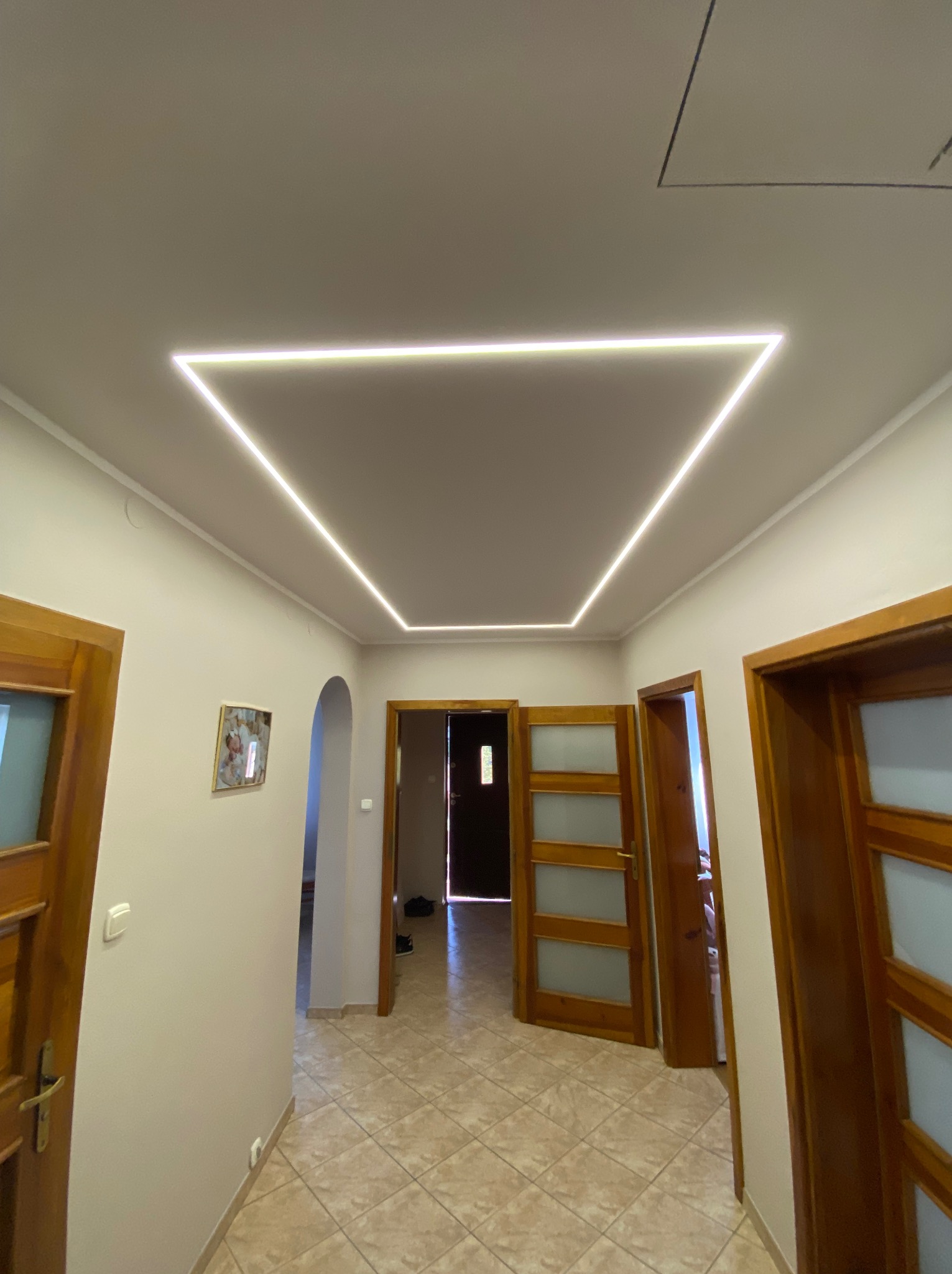 Korytarz z trzema parami drewnianych drzwi i oświetleniem LED w kształcie trapezu na suficie. Beżowe ściany i płytki podłogowe. Widok z dołu.