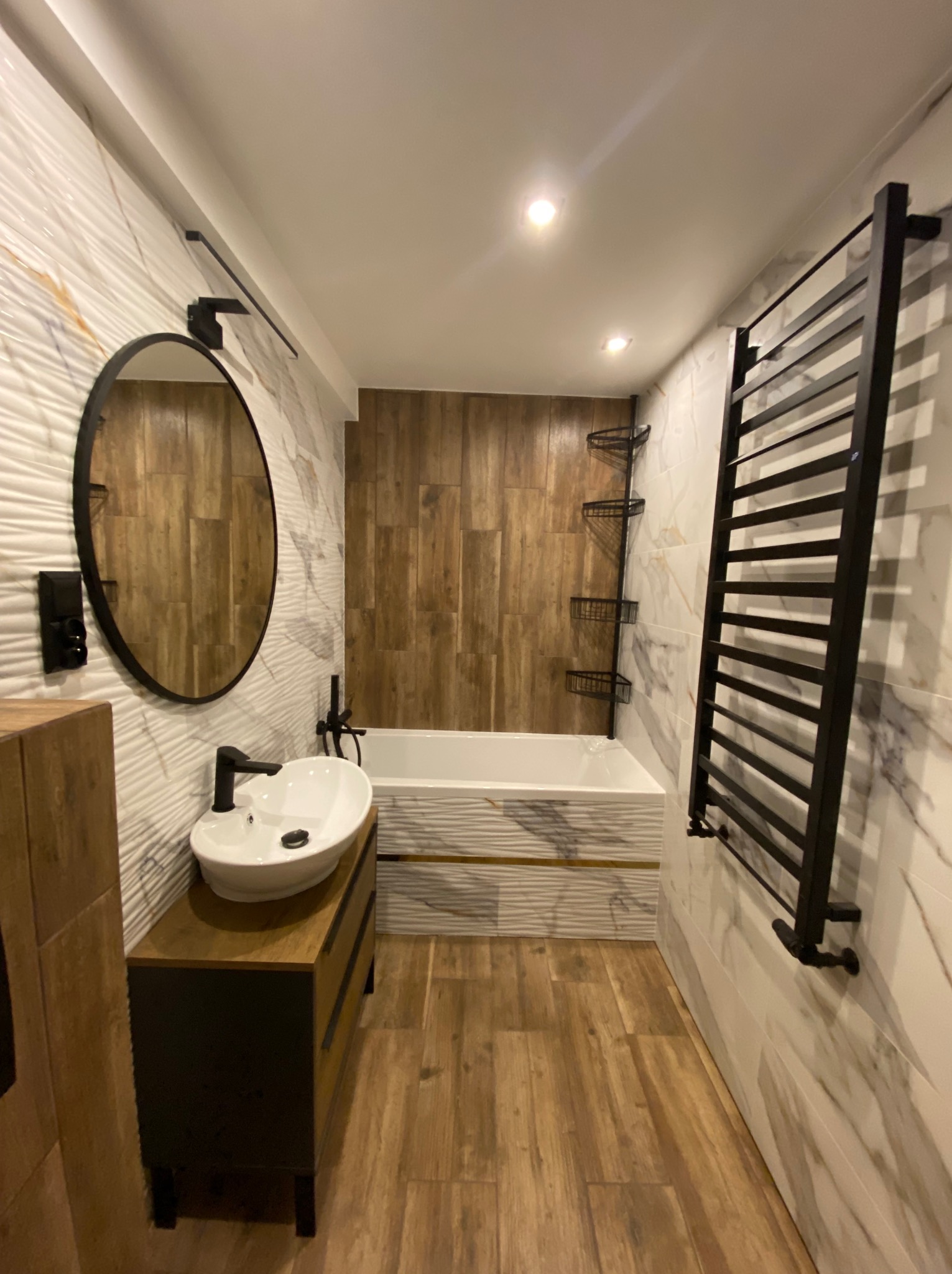 Nowoczesna łazienka po remoncie: drewnopodobne płytki, owalne lustro, biała umywalka nablatowa i czarny grzejnik drabinkowy. Wykończenie wnętrz w ciepłych barwach.