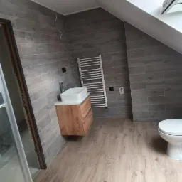 Wykończona łazienka na poddaszu z szarą okładziną ścienną imitującą drewno, drewnopodobną podłogą, białą umywalką na drewnianej szafce, grzejnikiem drabinkowym i toaletą.