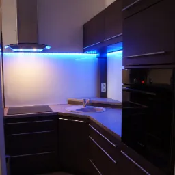 Nowoczesna kuchnia z ciemnymi, drewnopodobnymi frontami, podświetleniem LED w kolorze niebieskim i szklanym panelem nad blatem, widoczny piekarnik marki Bosch.