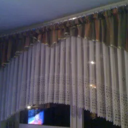 Zasłony woalowe z ażurowym wykończeniem i brązowo-beżowa krótka firana na karniszu, widoczne przez okno światło z telewizora.