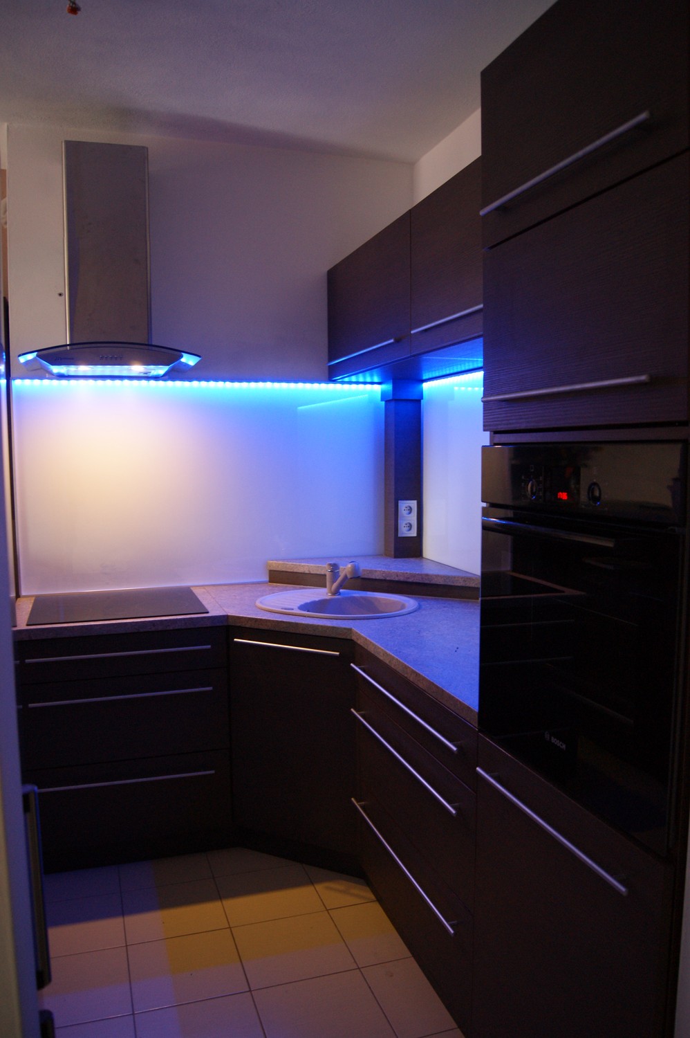 Nowoczesna kuchnia z ciemnymi, drewnopodobnymi frontami, podświetleniem LED w kolorze niebieskim i szklanym panelem nad blatem, widoczny piekarnik marki Bosch.