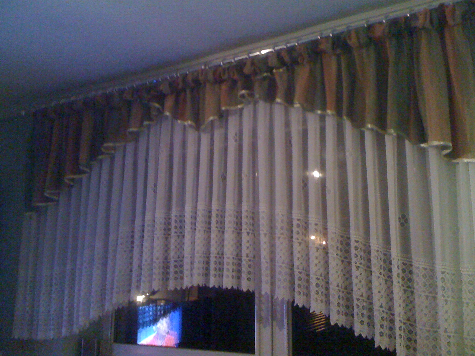 Zasłony woalowe z ażurowym wykończeniem i brązowo-beżowa krótka firana na karniszu, widoczne przez okno światło z telewizora.