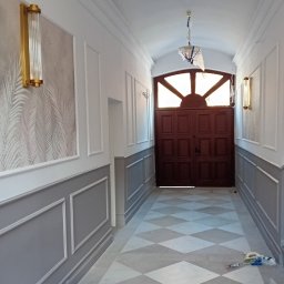 Kamil Kozuba - Korytarz z wykończonymi ścianami w stylu klasycznym, z szarą lamperią, białymi panelami i tapetą w liście, marmurowa podłoga w romby, złote kinkiety i drewniane drzwi wejściowe z łukowym nadświetlem.