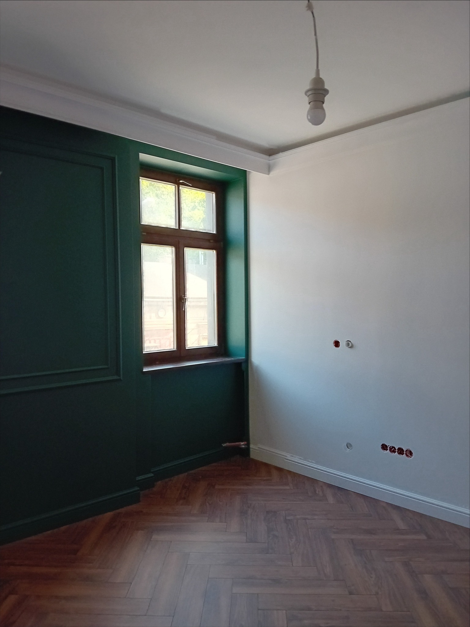 Wnętrze pokoju po remoncie: ściana pomalowana na ciemnozielono z dekoracyjnymi listwami, drewniane okno, parkiet ułożony w jodełkę, biała ściana z gniazdkami elektrycznymi i żarówka na kablu.