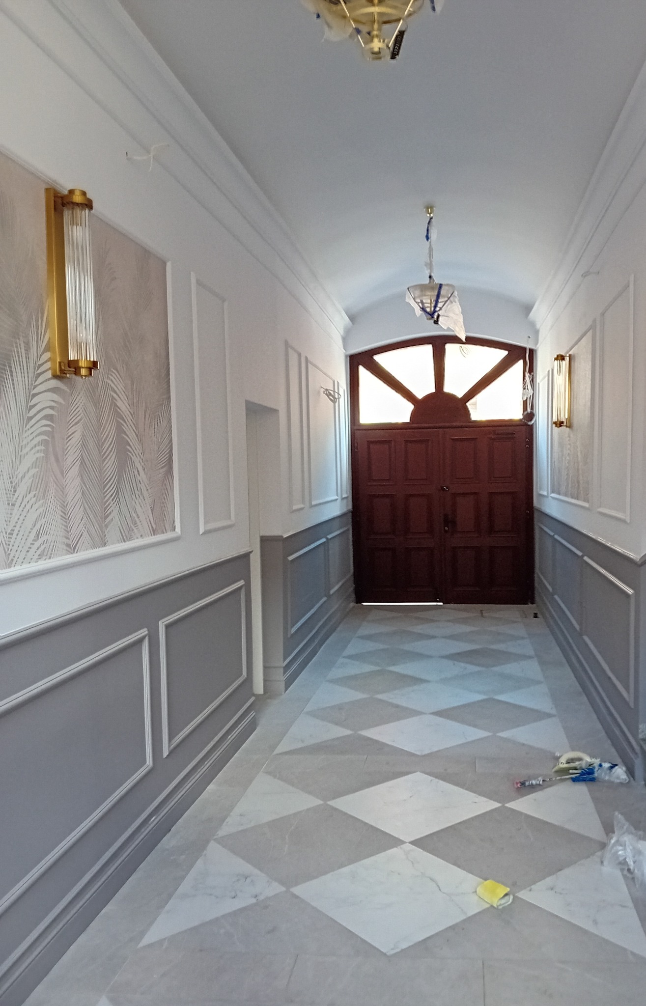 Korytarz z wykończonymi ścianami w stylu klasycznym, z szarą lamperią, białymi panelami i tapetą w liście, marmurowa podłoga w romby, złote kinkiety i drewniane drzwi wejściowe z łukowym nadświetlem.