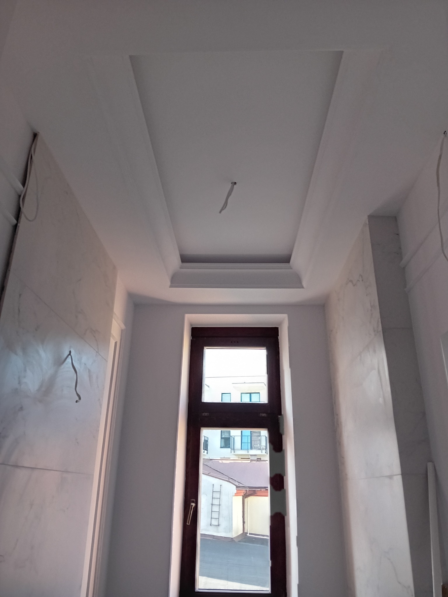 Wnętrze pokoju w trakcie prac wykończeniowych: ściany pokryte imitacją marmuru, sufit z dekoracyjnym obniżeniem i listwami, widoczne przewody elektryczne, okno z widokiem na budynki.