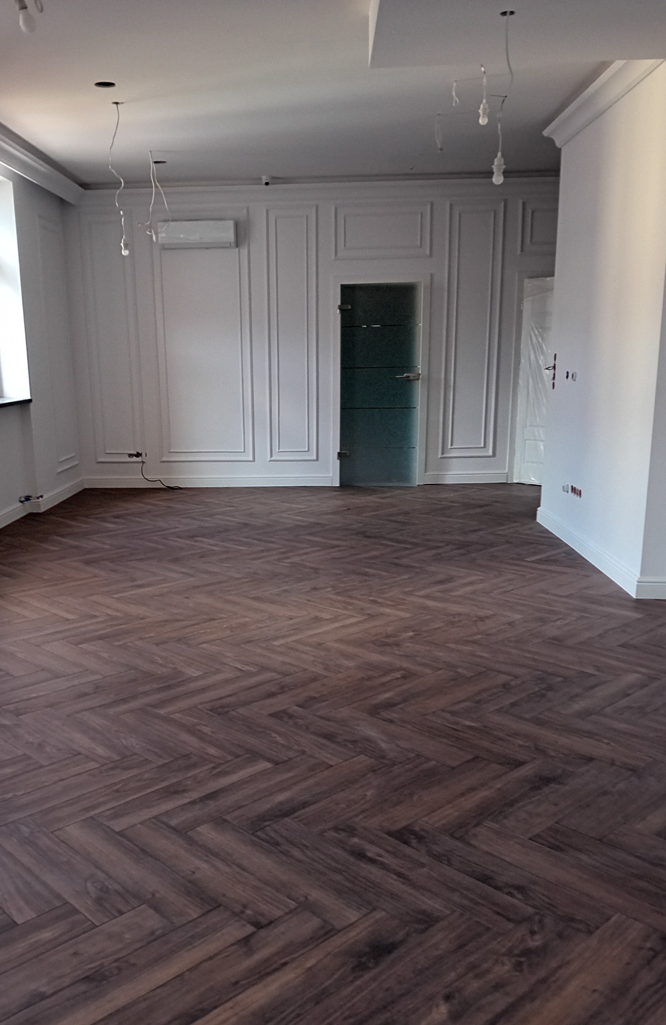 Wnętrze pokoju po remoncie z ułożonym parkietem w jodełkę, widoczne białe ściany z dekoracyjnymi listwami, szklane drzwi i niedokończone oświetlenie sufitowe.