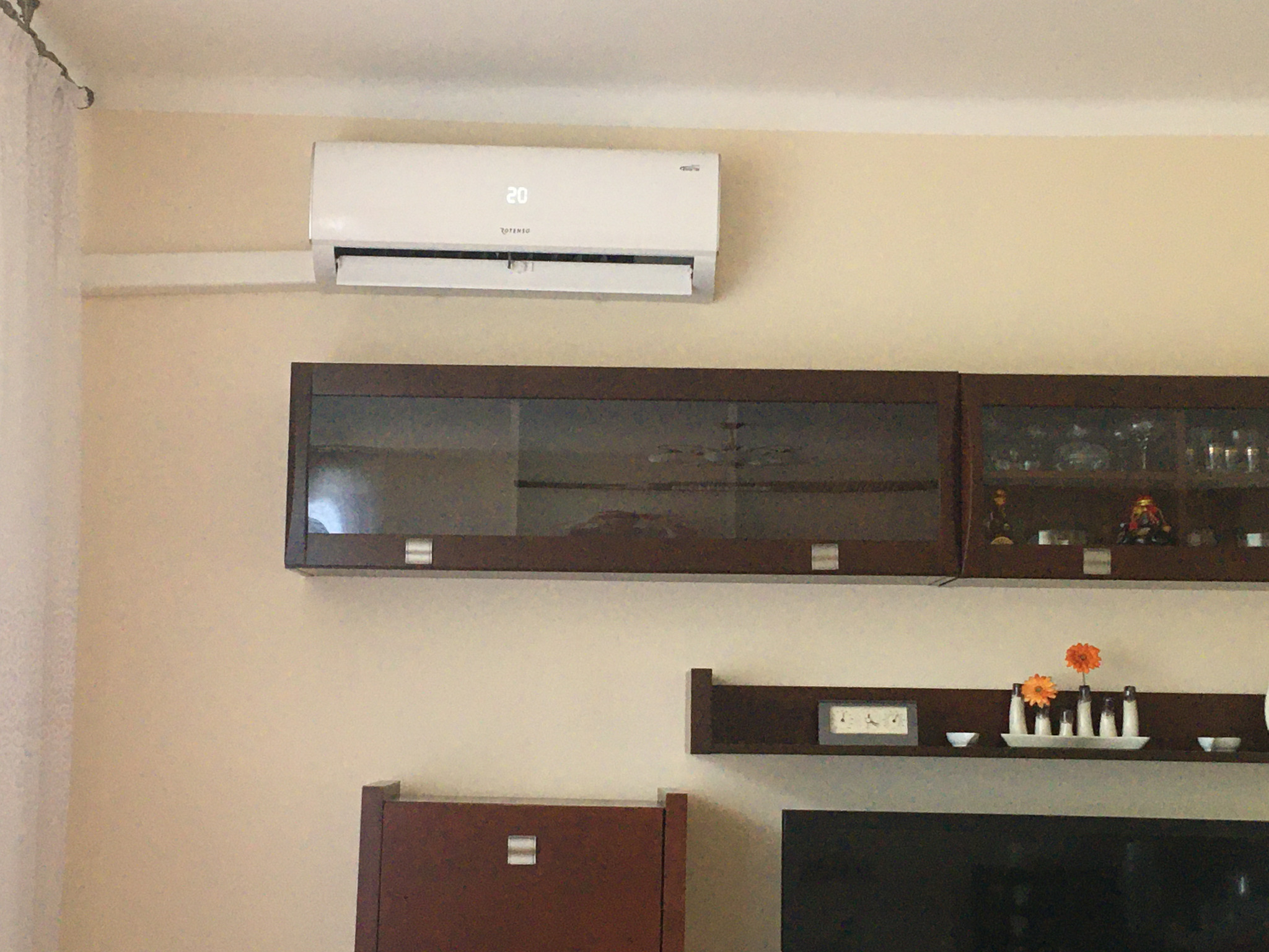 Biały klimatyzator ścienny z wyświetlaczem temperatury 20 stopni Celsjusza, zamontowany nad ciemnobrązowymi meblami w salonie.