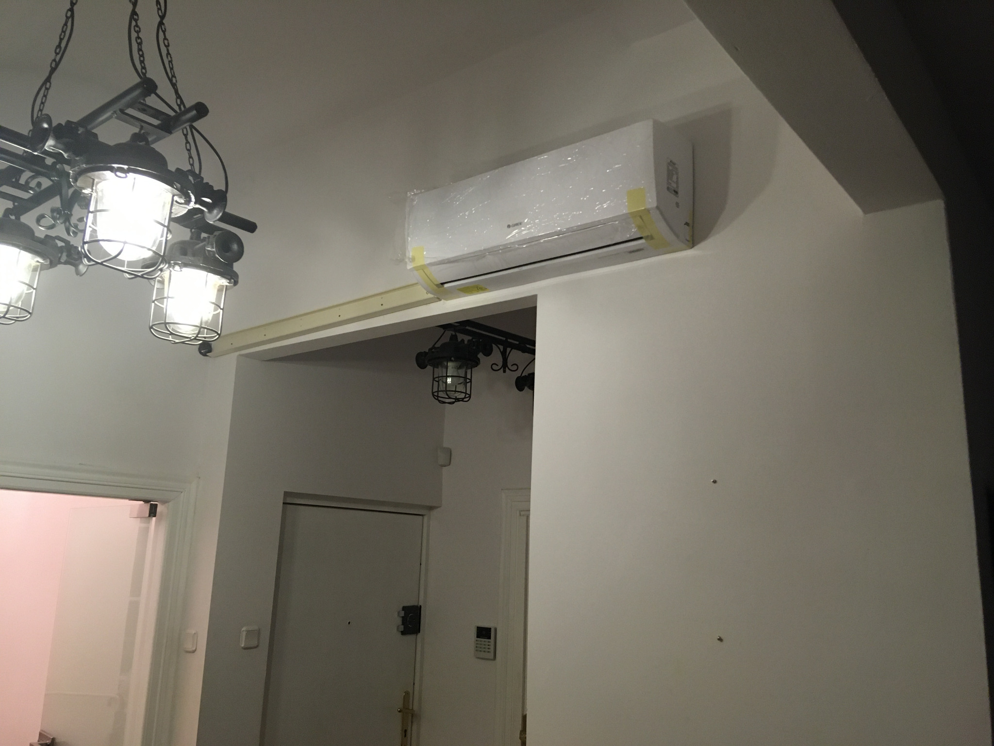 Nowa, biała jednostka wewnętrzna klimatyzatora, jeszcze w folii ochronnej z żółtymi taśmami, zamontowana pod sufitem w jasnym pomieszczeniu, widoczna industrialna lampa wisząca.