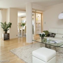 BUD MAX USŁUGI REMONTOWO BUDOWLANE ŁUKASZ CHRZAN - Jasne, przestronne wnętrze salonu z widokiem na jadalnię i kuchnię, z białymi meblami, szklanym stolikiem i puszystym dywanem w kolorze ecru.