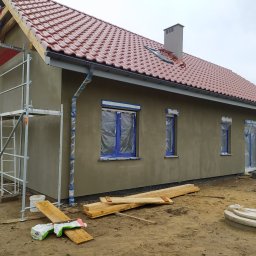 BUD MAX USŁUGI REMONTOWO BUDOWLANE ŁUKASZ CHRZAN - Dom jednorodzinny w trakcie nakładania tynku zewnętrznego, okna zabezpieczone folią, widoczne rusztowania i materiały budowlane, dach pokryty czerwoną dachówką ceramiczną.