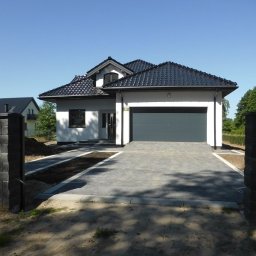 BUD MAX USŁUGI REMONTOWO BUDOWLANE ŁUKASZ CHRZAN - Nowoczesny dom jednorodzinny z szarą bramą garażową i brukowanym podjazdem, widok z poziomu gruntu, z elementami ogrodzenia z szarej kostki.