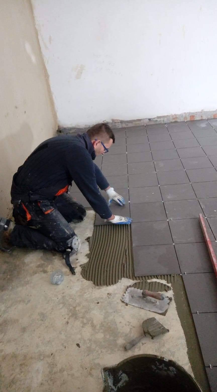 Pracownik w okularach układa ciemne płytki podłogowe, używając szpachelki do nałożenia kleju; widoczne narzędzia i krzyżyki dystansowe.